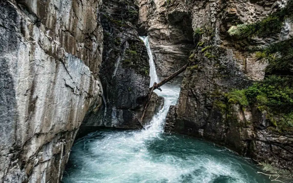 A cascading waterfall flows through a narrow rocky gorge, surrounded by mossy cliffs and vibrant greenery