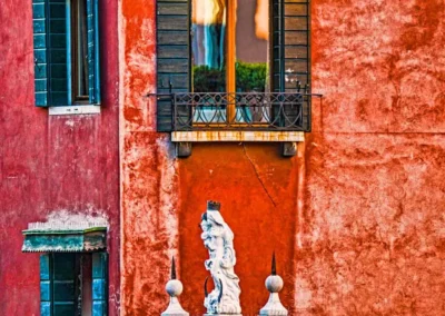 A decorative balcony with black shutters above a weathered red wall, featuring a white statue at the bottom center