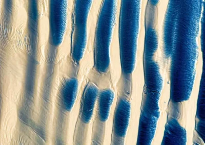 An aerial view of rippling sand and water, featuring contrasting blue waves against a light sandy beach background